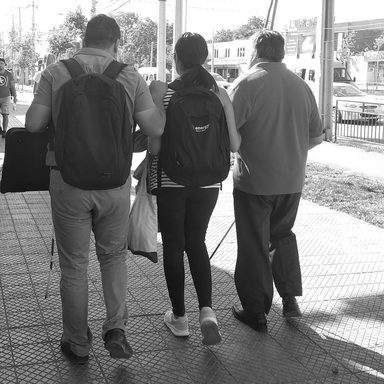 Tres personas caminan de espaldas por una vereda de Santiago. Dos llevan bastón blanco y la del centro va del brazo de los otros dos.