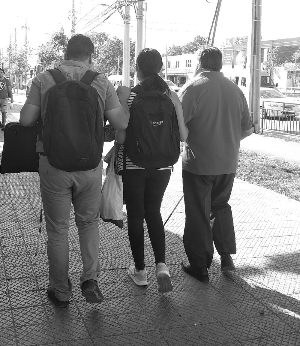 Tres personas caminan de espaldas por una vereda de Santiago. Dos llevan bastón blanco y la del centro va del brazo de los otros dos.